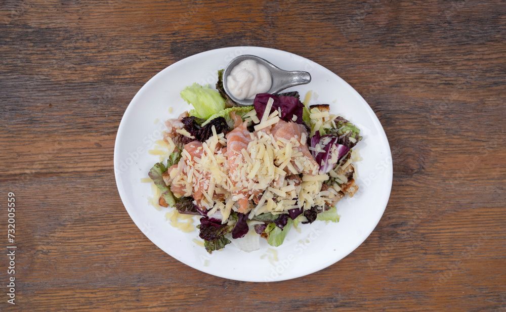 Top view of a Caesar salad with lettuce, parmesan cheese, bread croutons, salmon, bacon and Caesar dressing, in a white plate on the wooden table. Top view of a Caesar salad with lettuce, parmesan cheese, bread croutons, salmon, bacon and Caesar dressing, in a white plate on the wooden table.