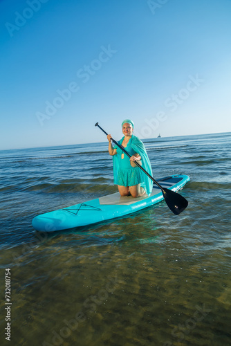 A woman in a turquoise swimsuit with a skirt and a scarf on her head on a SUP board near the seashore in the morning. © finist_4