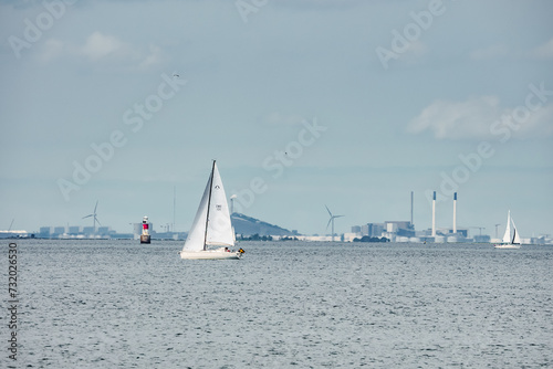 A serene seascape in the Oresund Strait with sailboat number 11 cruising, other boats nearby, and a clear sky. Wind turbines and industrial skyline hint at Copenhagen or Malmo.