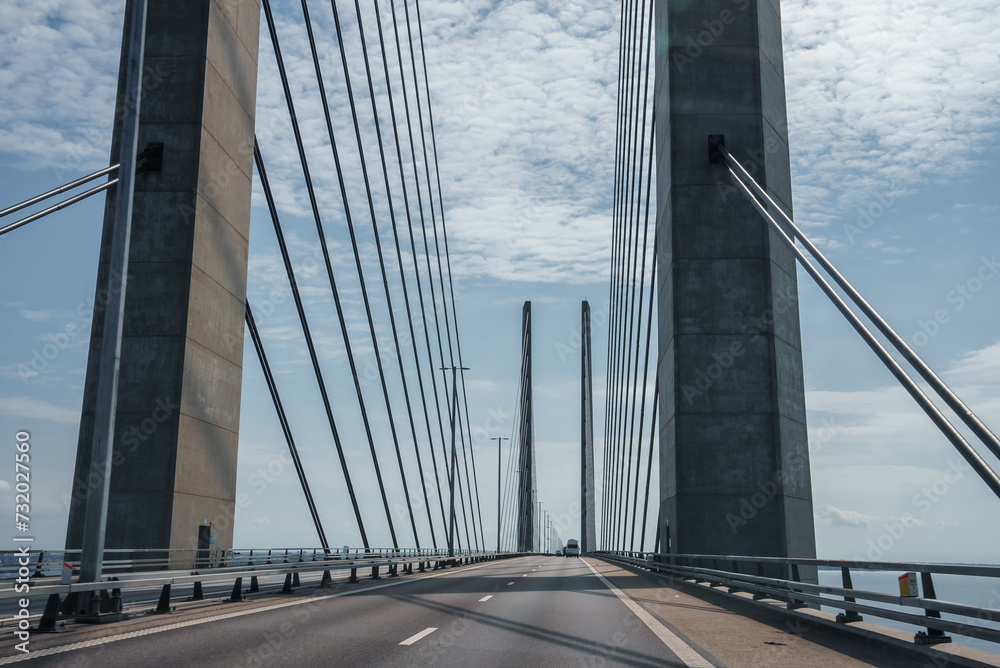 Capturing the sleek engineering of the Oresund Bridge, this image features its towering concrete supports and harplike cables under a clear blue sky, on a trafficfree multilane highway.