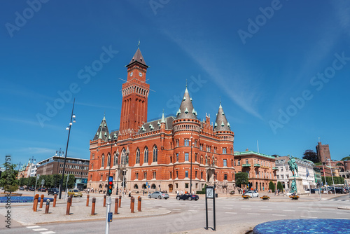 A sunlit red brick building with Romanesque and Gothic Revival elements, featuring a tower and arched windows, stands in a quiet plaza in Helsingborg. photo