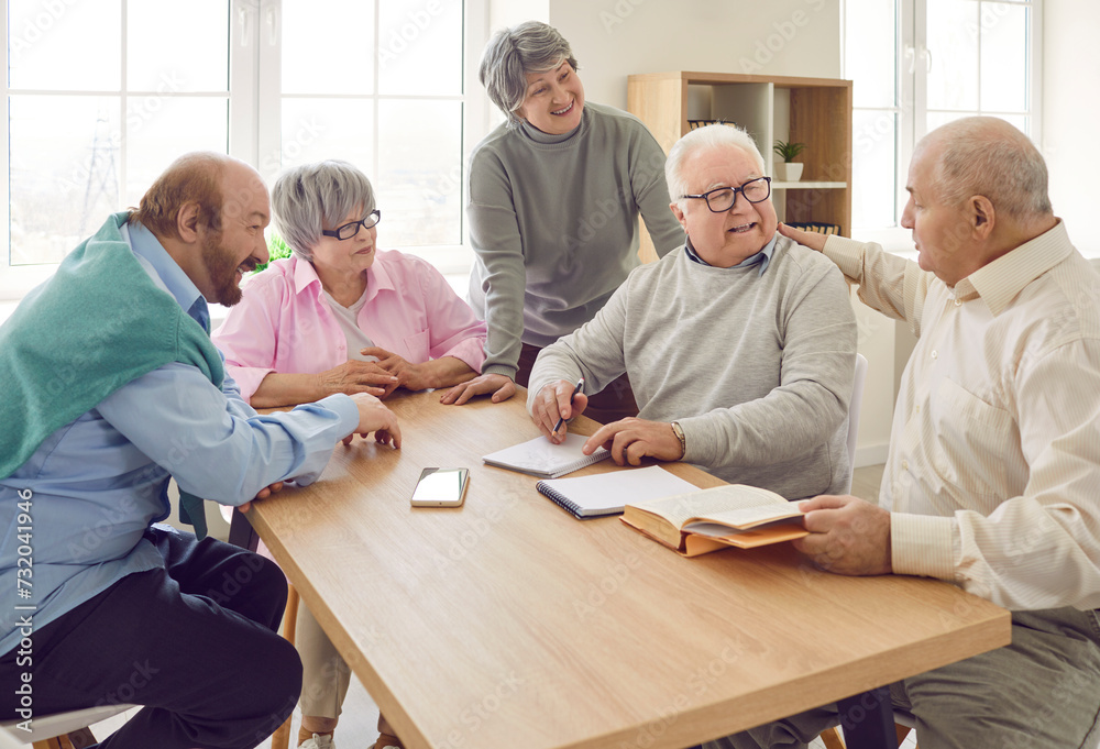 Group of cheerful elderly people interact with each other in modern nursing home. Elderly men and women are talking happily while sitting at table with phone, book and notebook. Old age concept. Group of cheerful elderly people interact with each other in modern nursing home. Elderly men and women are talking happily while sitting at table with phone, book and notebook. Old age concept.