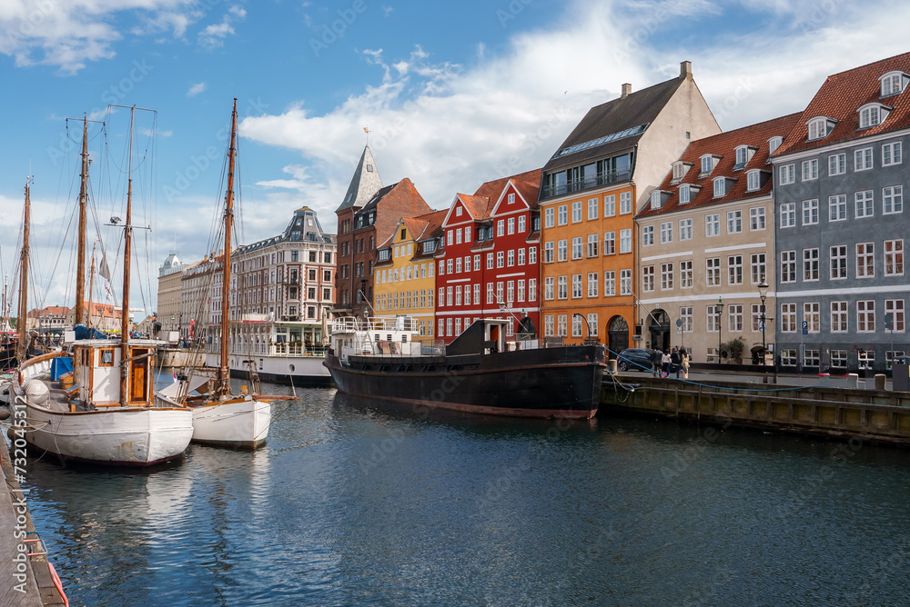 A serene day at Nyhavn, Copenhagen, with boats moored in the canal, reflecting the colorful facades of European townhouses, under a blue sky with scattered clouds.