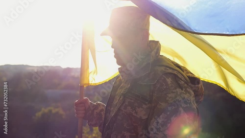 Soldier of ukrainian army stands with lifting blue-yellow banner at field. Male military in camouflage uniform holds a waving flag of Ukraine. Victory against russian aggression. Invasion resistance photo
