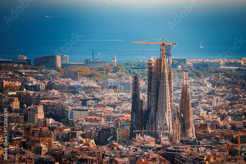Panoramic view of Barcelona featuring the unfinished Sagrada Familia with its spires and cranes, surrounded by the city's warmhued buildings and the Mediterranean Sea in the backdrop.