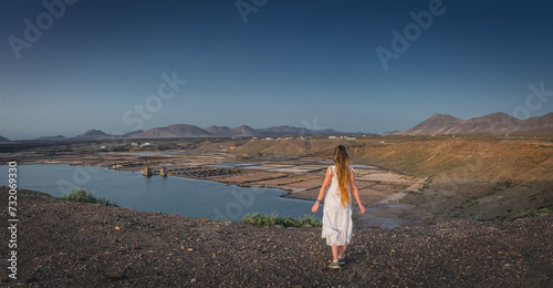 Girl in white dress stands back to camera on a cliff and looks on the Janubio flats (panoramic photo)