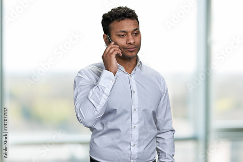 Confident young businessman wearing white shirt talking on transparent phone phone. Attractive company leader talks on futuristic phone on blur interior background.