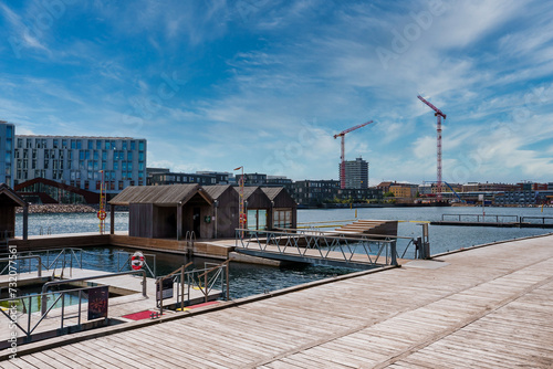 Serene waterfront, wooden boardwalk, and huts in Copenhagen. Calm bay, city skyline, cranes, clear skies show leisure and urban growth harmony.