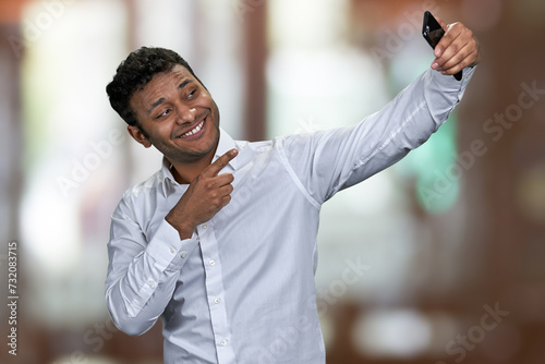 Joyful young man wearing white shirt gesturing index finger on camera while taking selfie with his smartphone. Abstract bokeh background.