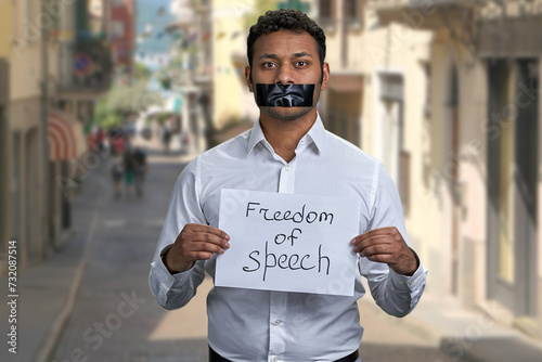 Young man with taped mouth holding paper card with inscription Freedom of speech. Blur narrow city street in the background. photo