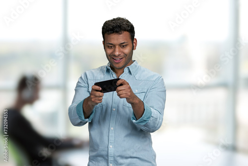 Young handsome man playing game on his smartphone. Office blur interior background.