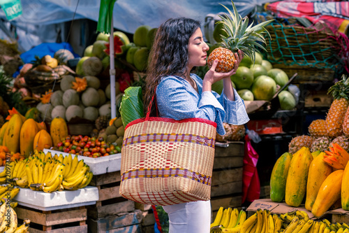 Beautiful Latin woman in the middle of a fresh fruit market, with a basket holding a pineapple in her hands.