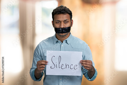 Dark-skinned censored man holding paper banner with inscription Silcence. Interior blur background.