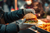 A man of a street food seller handing over a delicious burger that he has in his hand, wrapped in paper from a kiosk at a street market, at an open-air festival
