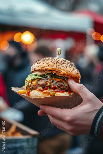 Person in fast food street market, holding in hand delicious burger in paper wrap, freshly made cheeseburger, outdoors, closeup. Summer