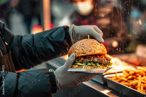 A man of a street food seller handing over a delicious burger that he has in his hand, wrapped in paper from a kiosk at a street market, at an open-air festival photo