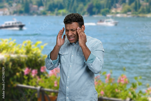 Stressed young man suffering from terrible headache standing outdoors. Man with headache on sea background.