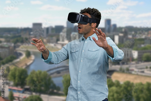 Young shocked man wearing vr glasses outdoor. Blur city skyscrapers in the background. Scared surprised man wearing vr headset.