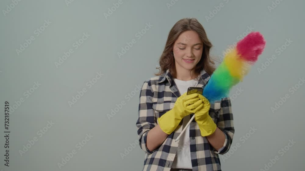 A Happy Young Woman in Yellow Gloves and With a Dustpan, Rejoices at Success on the Phone on the Gray Background.Wireless and App-controlled Cleaning Appliances. A Happy Young Woman in Yellow Gloves and With a Dustpan, Rejoices at Success on the Phone on the Gray Background.Wireless and App-controlled Cleaning Appliances.