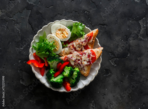 Delicious diet lunch, breakfast - boiled egg, fresh vegetable salad, broccoli and hot sandwiches with tomato and cheese on a dark background, top view