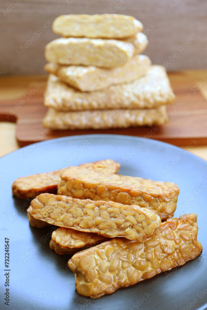Plate of pan fried Tempeh with Stack of the Uncooked in the Backdrop Plate of pan fried Tempeh with Stack of the Uncooked in the Backdrop