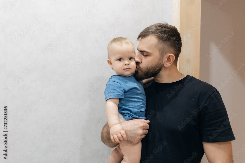 Father hugs baby boy, young man kisses his son on head in living room. Concept lifestyle parenting fatherhood moment