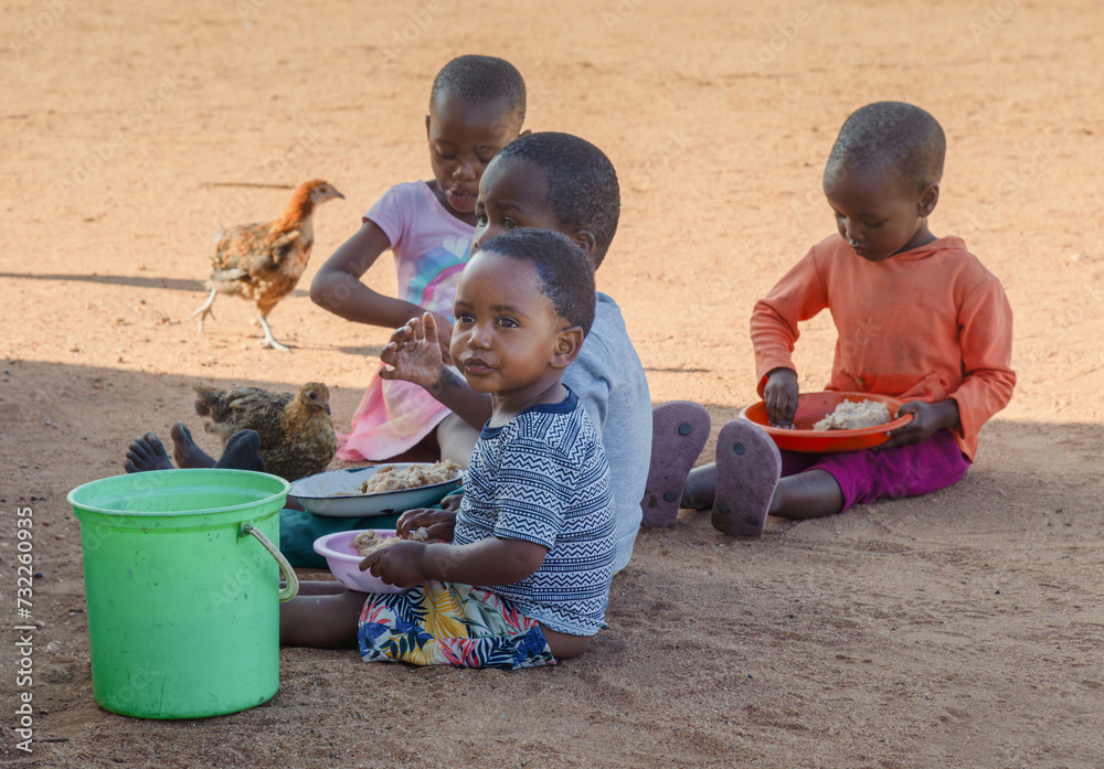 hungry village african children in the yard , sited on the ground eating staple food from plastic plates hungry village african children in the yard , sited on the ground eating staple food from plastic plates
