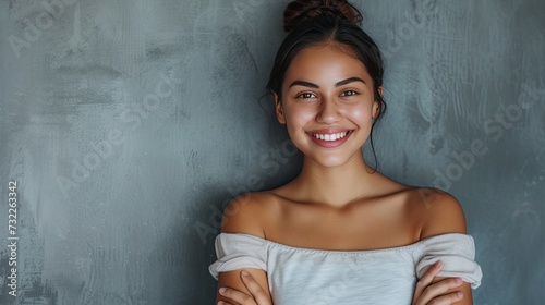 Portrait of a young latin woman with pleasant smile and crossed arms isolated on grey wall with copy space. Beautiful girl with folded arms looking at camera against grey wall - generative ai © Nia™