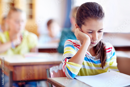Child, school and desk with book or bored in classroom or education lesson or reading, knowledge or studying. Girl, kids and unhappy or learning or tired pupil with paper or fatigue, student or moody photo