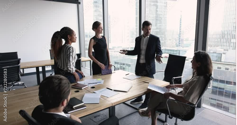 Business leader man talking to diverse team of managers sitting and standing at meeting table, discussing teamwork, cooperation, tasks, project management, instructing colleagues Business leader man talking to diverse team of managers sitting and standing at meeting table, discussing teamwork, cooperation, tasks, project management, instructing colleagues
