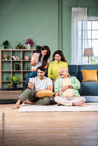 Happy smiling indian senior parents with adult son using smartphone while sitting on sofa at home.