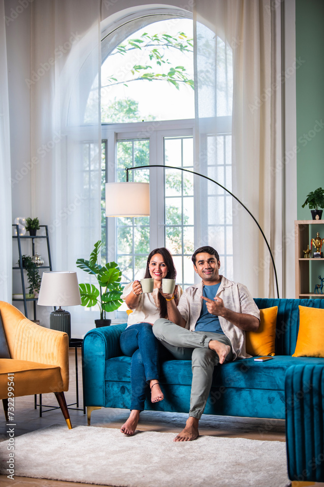 Fototapeta premium Cheerful young beautiful Indian couple having coffee together on sofa at home..