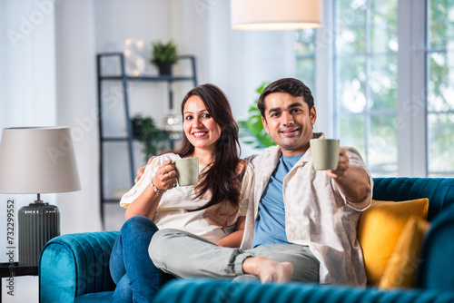 Cheerful young beautiful Indian couple having coffee together on sofa at home..