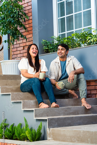 Cheerful young beautiful Indian couple having coffee together on stairs at home..