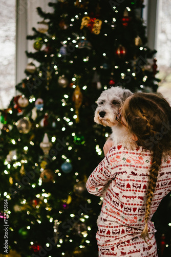 Girl in Christmas pajamas gives white puppy hug in front of tree photo