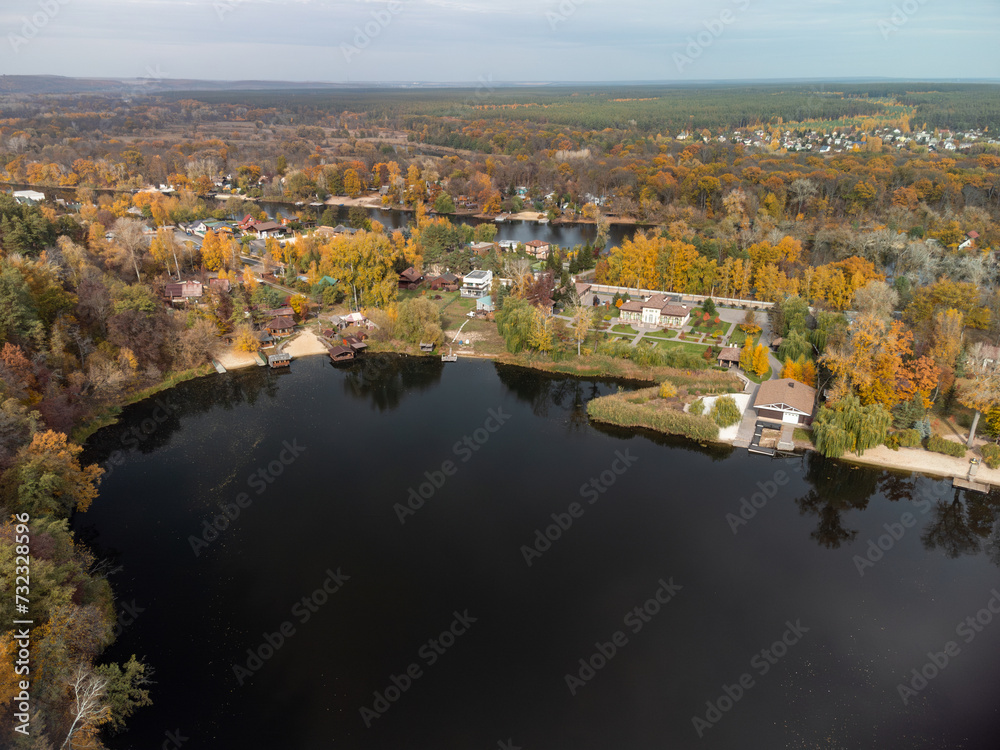 Aerial autumn cottages on river with colorful forest. Flying above vibrant autumnal village in Ukraine