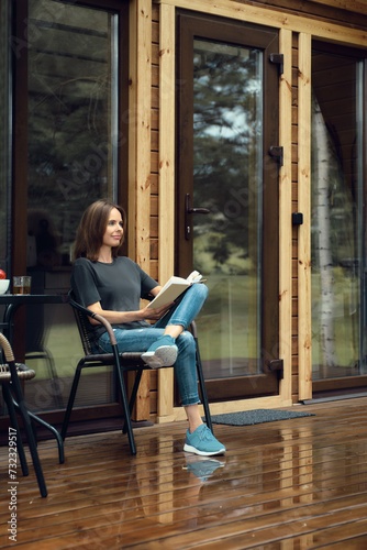 Cute woman in casual clothing sits reading on a wet terrace of a log cabin after the rain, enjoying fresh air