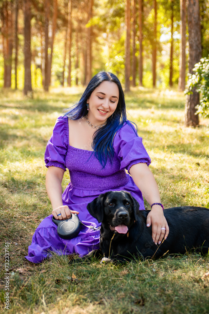 A young woman in a purple dress sits next to a black Labrador retriever against the backdrop of the park. A young woman in a purple dress sits next to a black Labrador retriever against the backdrop of the park.