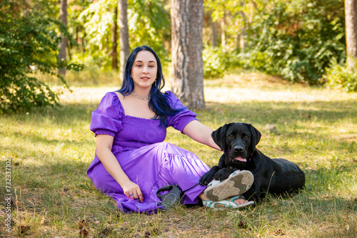 A young woman in a purple dress sits next to a black Labrador retriever against the backdrop of the park. © Михаил Гута
