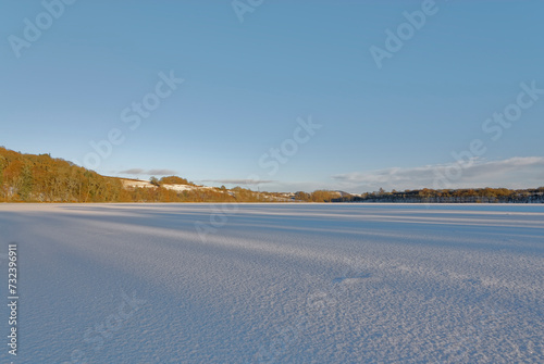 A Frozen and Snow covered Loch Clunie with long shadows across the Loch surface on a cold day in January.