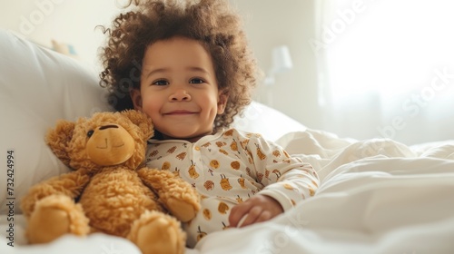 A joyful child with curly hair wearing a white onesie with yellow patterns sitting on a bed with a brown teddy bear.