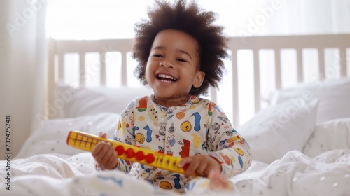 A joyful child with curly hair wearing colorful pajamas sitting on a bed and holding a toy smiling brightly.