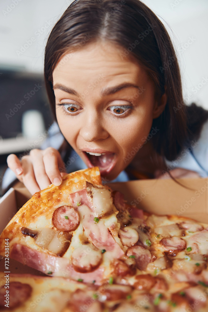 Hungry woman holding pizza box with mouth wide open, eagerly reaching for a slice of pizza in front of her