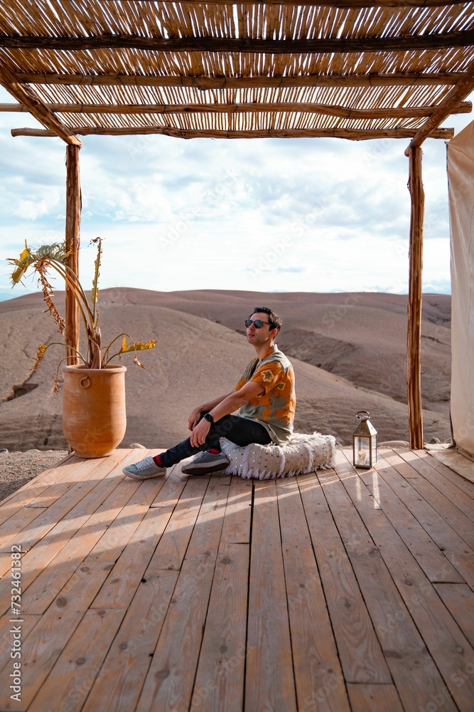 Man with sunglasses sitting on a wooden deck in a sunny outdoor setting. Agafay desert, Morocco.
