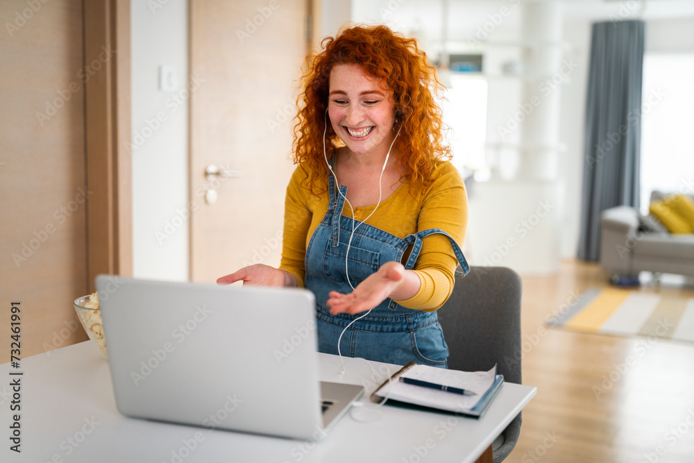 Young adult female college student having a video call on her laptop. Woman with red hair and headphones laughing while sitting indoors. Young adult female college student having a video call on her laptop. Woman with red hair and headphones laughing while sitting indoors.