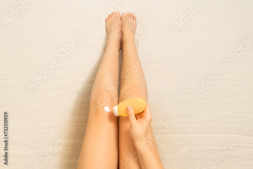 Top view on slim woman legs on sandy beach. Female applying sunscreen cream for protection skin sitting and sunbathing on sand. Relaxation at summer vacation. Aerial view.