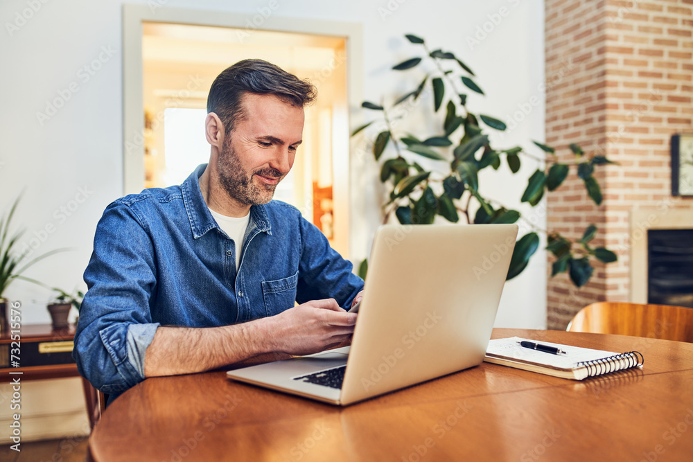 Smiling adult man using mobile phone and laptop sitting by table at home Smiling adult man using mobile phone and laptop sitting by table at home