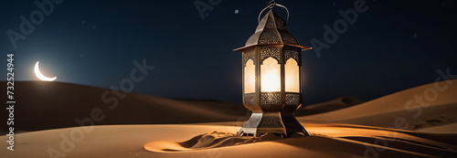 Panoramic banner with traditional ornamental glowing Arabic lantern on the sand in the middle of the desert on the background of night sky and crescent moon. Header with copy space for Ramadan Kareem