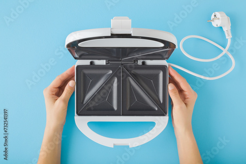 Murais de parede Woman hands holding white opened plastic sandwich maker with dark black nonstick surface on blue table background