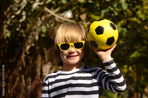 Cute little preschooler boy holding a yellow soccer ball. A child in sunglasses holds a ball, laughs